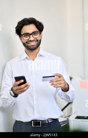Smiling indian business man using phone holding credit card making e payment. Stock Photo