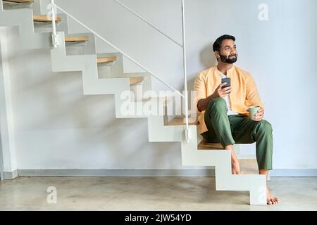 Indian man sitting at home sitting on stairs wearing earbuds using mobile phone. Stock Photo
