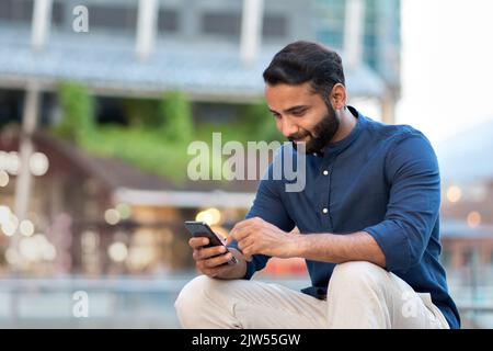 Smiling indian businessman using smartphone sitting on urban city street. Stock Photo