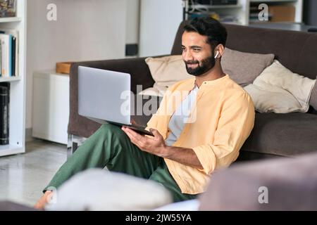 Happy indian man sitting at home wearing earbud using laptop computer. Stock Photo