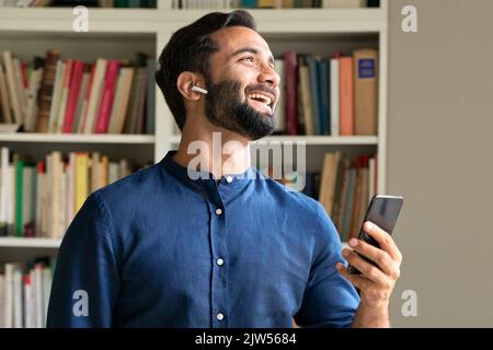 Happy indian business man at work wearing earbud using smartphone. Stock Photo