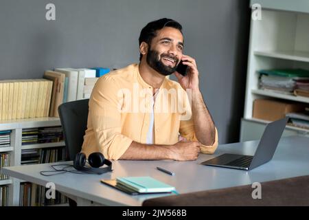 Happy indian business man sitting at work desk talking on phone making call. Stock Photo