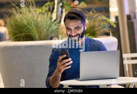 Happy indian business man using smartphone working on laptop at night outside. Stock Photo