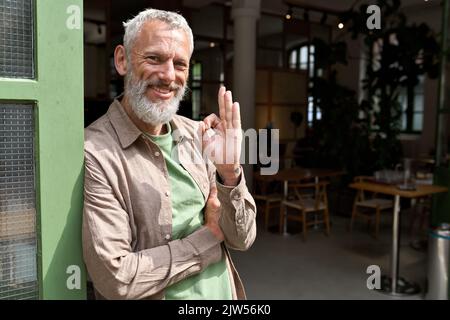 Happy older man small business owner showing ok sign standing outside cafe. Stock Photo