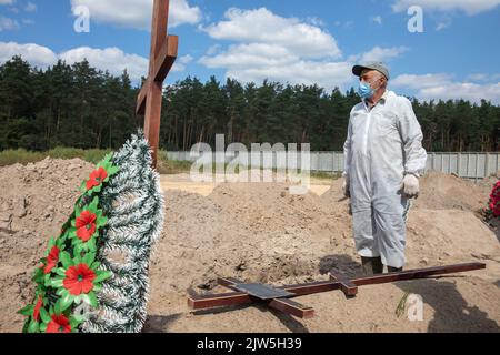 A man in a protective suit places a cross on a grave. Coffins containing the bodies of unidentified persons killed in Bucha district at the time of the Russian occupation, during a burial ceremony at a cemetery in Bucha, Kyiv region, Ukraine.. Months after the withdrawal of the Russian forces from the Bucha district, those bodies that were not identified have been buried and marked with numbers at a cemetery in Bucha. Stock Photo