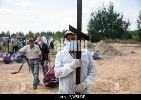 A man in a protective suit and face mask carries a funeral cross to be installed on a fresh grave. Coffins containing the bodies of unidentified persons killed in Bucha district at the time of the Russian occupation, during a burial ceremony at a cemetery in Bucha, Kyiv region, Ukraine.. Months after the withdrawal of the Russian forces from the Bucha district, those bodies that were not identified have been buried and marked with numbers at a cemetery in Bucha. Stock Photo