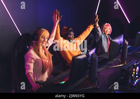 Three teenagers wearing colorful clothes and headsets celebrating victory in online with high fives. Professional gaming tournament. Color neon lights. Indoor shot. High quality photo Stock Photo