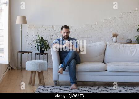 Serious single man using modern cellphone seated on sofa Stock Photo