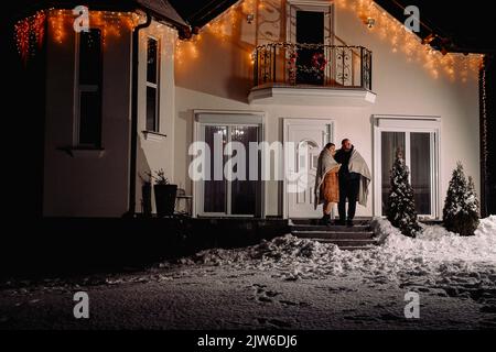 Full-length photo of a happy couple standing in front of their house on winter evening in warm clothes covered with blanket. Stock Photo