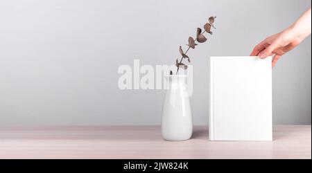 Banner with hand putting book mockup near vase with flowers at table. Minimalist style. Reading leisure, harmony concept. Novel with white cover. Copy Stock Photo