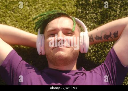 Portrait of young man in wireless headphones listening to music, closing eyes from bright sun, lying on lawn Stock Photo