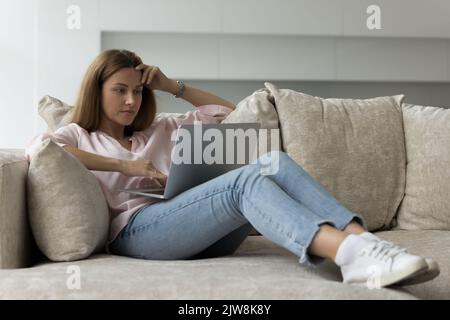 Focused thoughtful freelance business woman using laptop on home sofa Stock Photo