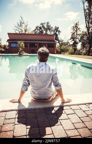 Young man seen from behind is sitting by the pool. Businessman takes a relaxing moment by the pool. Stock Photo