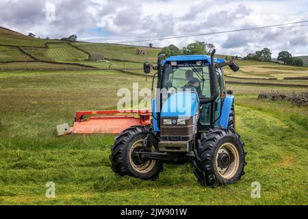 Close up of a farmer in a blue tractor cutting the grass for silage in fields near Appletreewick in the Yorkshire Dales National Park, England, UK Stock Photo