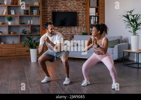 Cheerful young african american husband and wife in sportswear doing squat exercises Stock Photo