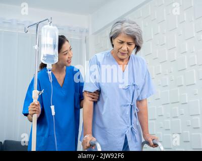 Elderly Asian woman patient trying to walk on walking frame held and carefully supported in arms by caregiver, young polite female assistant nurse in Stock Photo