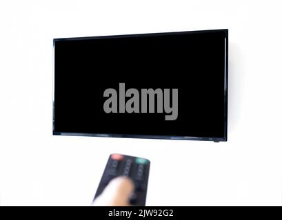 View of mockup black blank space on TV screen isolated on white wall background with blurred focus of remote control in person hand on foreground. Emp Stock Photo