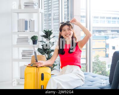 Young pretty happy Asian woman touching sunglasses on head holding passport with smiling, sit on sofa near beach hat, yellow suitcase in white living Stock Photo