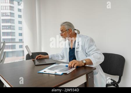 Senior healthcare worker doing some paperwork and using laptop while working at doctor's office Stock Photo