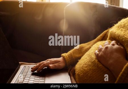 Pregnant woman in a cosy sweater working on laptop in the morning during sunrise. Beautiful golden sunrise with lens flare and sunlight shining. Stock Photo