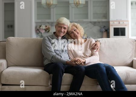 Happy cute senior married couple sitting on home sofa, hugging Stock Photo