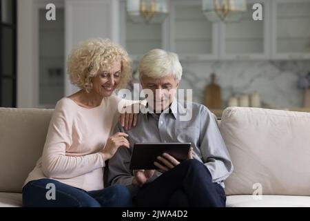 Happy senior couple using online app on tablet computer, laughing Stock Photo
