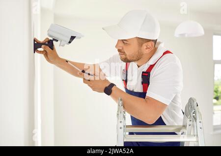 Electrician worker installs and screws surveillance video camera on wall in bright office space. Stock Photo