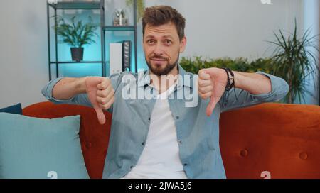 Dislike. Upset man showing thumbs down sign gesture, expressing discontent, disapproval, dissatisfied bad work at modern home apartment indoors. Displeased young guy in living room sitting on sofa Stock Photo