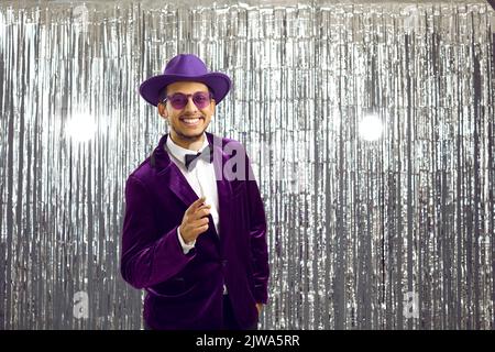 Happy man in purple suit, hat and glasses presents something on blank background Stock Photo
