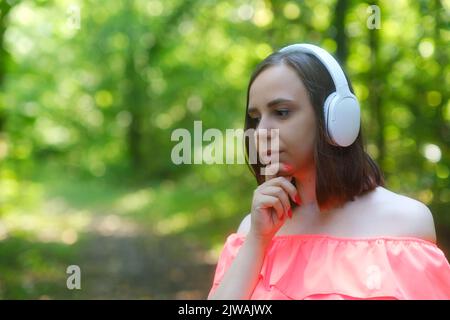 Portrait of young beautiful woman in wireless white headphones listening to music, scratching chin, looking down thoughtfully, standing in forest Stock Photo