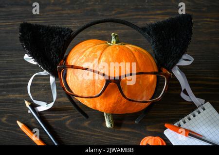 Halloween still life, happy pumpkin with glasses on wooden table, school supplies on Halloween. Orange pumpkin, notepad, pens, pencil on dark wood. Co Stock Photo