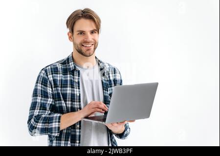 Positive handsome confident caucasian stylish young man, freelancer, student, programmer, standing on a white isolated background, holds an open laptop, looking to the camera, smiling friendly Stock Photo