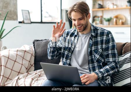 Distant communication concept. Positive friendly handsome caucasian stylish young man, sitting on a sofa in living room, talking by video conference using laptop, waves hand, smiling happily Stock Photo