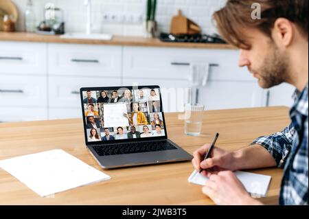Online education. Side view of a smart caucasian male student and group of multiracial people on a laptop screen. Clever guy listening to an online lecture, takes notes, gain knowledge distantly Stock Photo