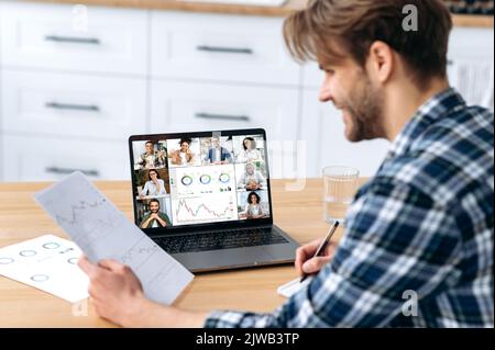 Side view of caucasian clever male top manager, sits in kitchen at home talking by video call with group of multiracial successful coworkers discuss company promotion, financial strategy, new project Stock Photo
