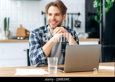 Photo of a successful modern caucasian male freelancer, stylishly dressed, sitting at desk with laptop in a kitchen at home, working on a creative project, looking to the side, smiling happily Stock Photo