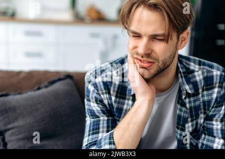 Problems with teeth. Toothache. Upset caucasian guy is sitting on the couch at home, holding his hand to his cheek, having a toothache, needs a dentist consultation, sad facial expression Stock Photo