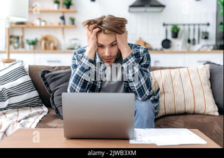 Confused upset caucasian young stylish man, sitting on sofa in living room, working online, frustrated looking at laptop screen, failed project, failed deal, holding hands on head, despair emotions Stock Photo