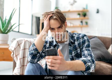 Sad confused upset caucasian young stylish man, sitting on a floor near a sofa in living room, frustrated looking at smartphone screen, failed project, got bad news or message, despair emotions Stock Photo