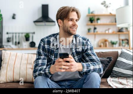 Positive handsome stylish caucasian young man, sitting with smartphone in hands on couch at home in living room, messaging online with friends, browsing websites, looks away, dreaming, smiling Stock Photo