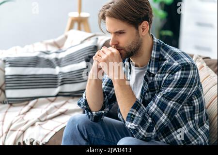Sad depressed stressed caucasian man, sitting in the living room on the couch, looking ahead of himself, experiencing anxiety due to personal problems, unfulfilled expectations, something hurts Stock Photo