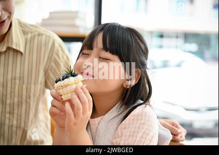 Happy cute Asian little girl enjoys eating cake or dessert at the cafe coffee shop with her dad. Stock Photo