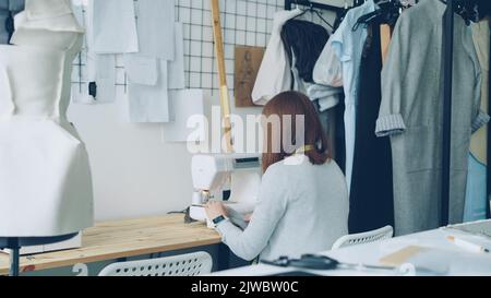 Back view of young woman clothing designer working with sewing machine sitting at studio desk. Sewing items, tailoring dummy and half-finished women's clothes are visible. Stock Photo