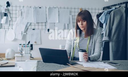 Young attractive seamstress is working with laptop and drinking take-out coffee in her tailoring studio. She is having rest after work. Relaxation in workplace concept. Stock Photo