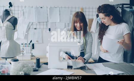Young woman is working with sewing machine and checking stitches when her colleague is coming to her with sketch. Women are looking at fabric and talking. Teamwork in fashion industry concept. Stock Photo