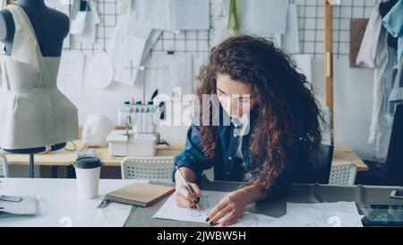 Young female artist fashion designer is drawing women's garment sketch with pencil at table in modern studio. Mannequin, sewing items, take-away coffee are visible. Stock Photo
