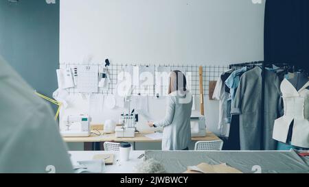 Back view of creative female tailor taking sketches from tailoring desk and hanging them on white wall above table. Planning, designing and creating new collection concept. Stock Photo