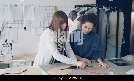 Team of creative designers is working with clothing patterns and fabric in tailor's shop. Young women are concentrated on measuring. Multiple sewing items and tools in background. Stock Photo
