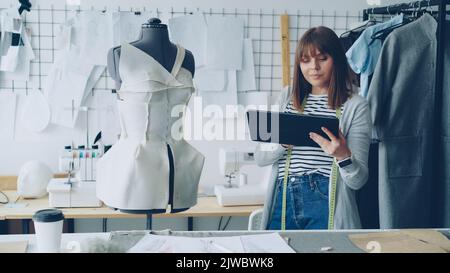 Beautiful female tailor is turning tailoring dummy to check pinned clothing and working with tablet to enter information. Using modern technologies in clothes manufacturing concept. Stock Photo