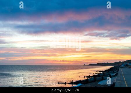 The dawn sky over the sea on the Kent coast at Reculver with the twin towers of the 12th century church silhouetted in the distance against an orange sky. In the foreground the silhouetted breakwaters, groynes, and beach at Henre Bay. Thick cloud overhead underlit by orange light. Stock Photo